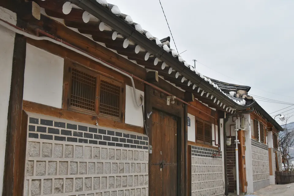 나무 창살 창과 돌담, 끝이 흰 기와가 이어지는 한옥의 옆모습